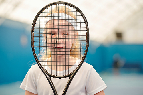Little blond girl in white t-shirt and headband holding tennis racket in front of face and looking at you through net in modern stadium