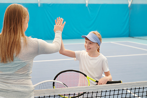 Happy teenage girl in activewear holding tennis racket while giving high five to her trainer over net before new training on modern stadium