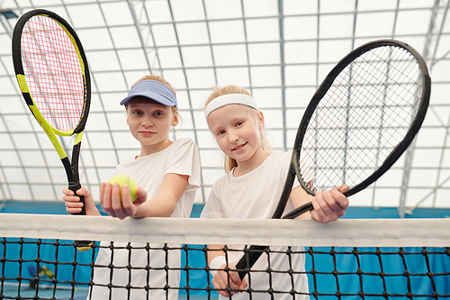 Two cute blond girls in white sportswear standing by net at stadium in front of camera and holding tennis rackets ready for play or training