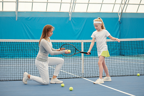 Female tennis instructor in activewear holding ball close to racket net while standing on knee in front of blond girl and consullting her on stadium