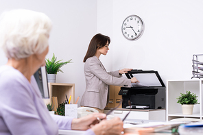 Young elegant office secretary or insurance agent making copy of document for senior client while standing by xerox machine