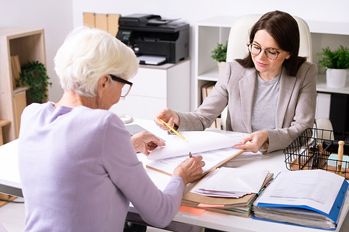 Friendly financial adviser working with senior woman and showing place for signature in document