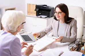 Smiling attractive young woman in glasses sitting at desk and pointing at paper while explaining senior lady how to fill form