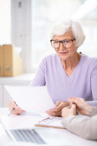 Concentrated elderly woman in glasses sitting at table and reading document before signing