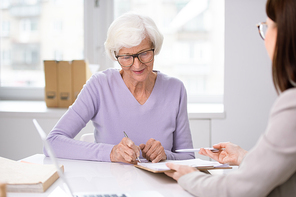 Senior client putting her signature in insurance agreement after discussion of its terms and conditions with agent at meeting