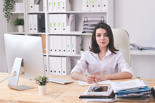 Portrait of serious attractive young businesswoman in white blouse making notes in diary in modern office