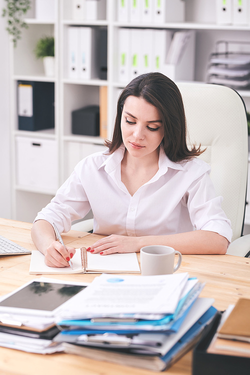 Pretty young serious female in white shirt sitting in armchair by desk and making working plan in notebook on background of shelves