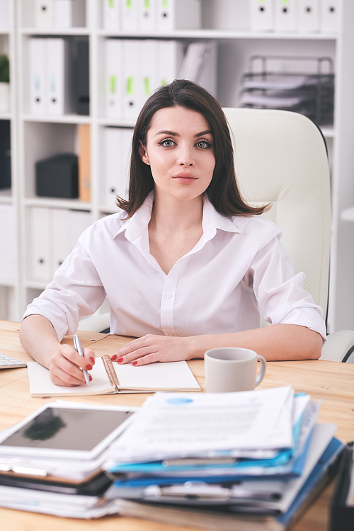 Portrait of content female office manager sitting at desk and writing in organizer