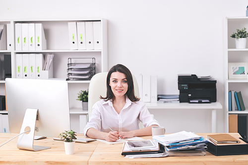Young successful businesswoman in formalwear sitting by desk in office, making working plan or notes about new business project