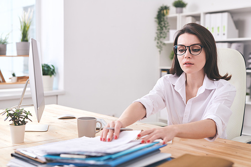 Attractive young businesswoman sitting at desk in office and finding documents in stack