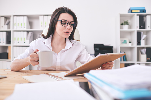 Busy young office woman sitting at table and reading papers while drinking coffee in office