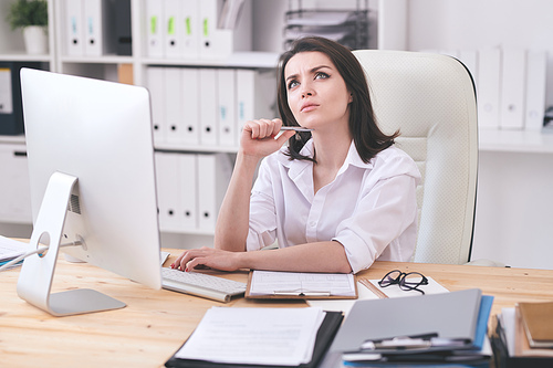 Pensive young business lady in white blouse sitting at table in office and thinking about online project