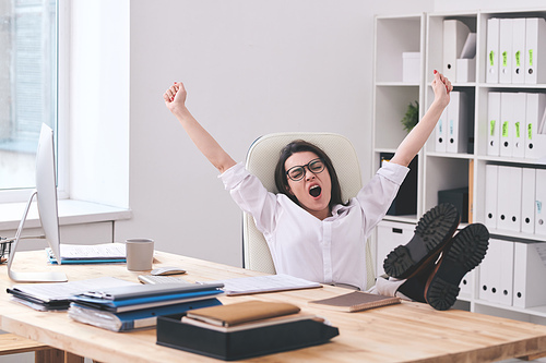 Tired young office woman sitting with feet on table and yawning with outstretched arms in office