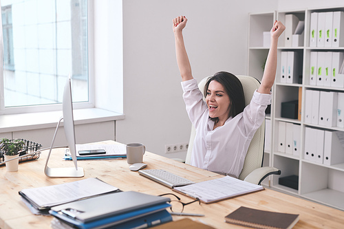 Positive emotional young businesswoman excited about success of project raising hands up while looking at computer monitor