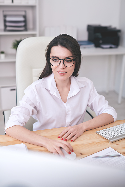 Smiling young smart lady in glasses sitting at desk and using modern computer in office