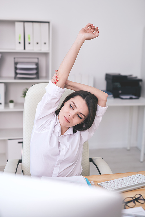 Calm attractive young businesswoman in white shirt stretching at desk while taking break at workplace
