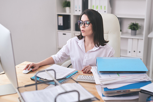 Busy young woman in blouse sitting at desk and using computer while working on pile of documents