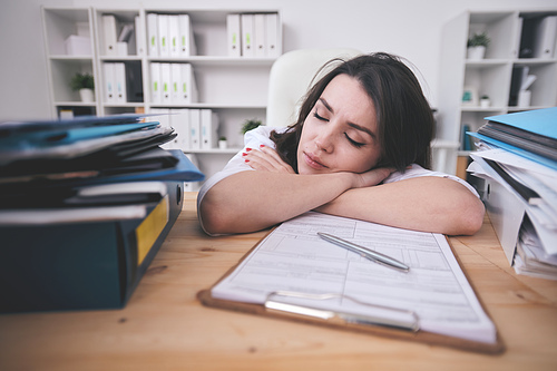 Tired young female office manager or accountant napping on desk in front of clipboard with financial papers after hard working day