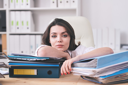 Portrait of young businesswoman exhausted with paperwork leaning on folders with work papers in office