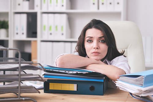 Young unhappy female accountant or secretary lying on thick stack of financial papers while sitting by desk in front of camera