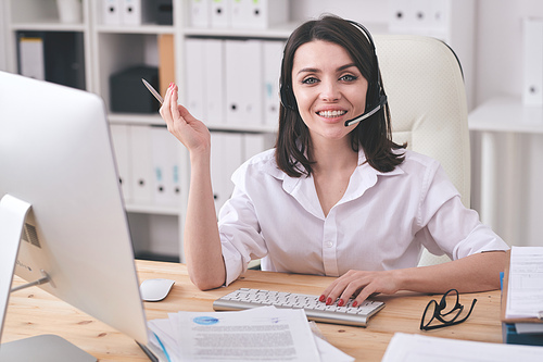 Young smiling customer support representative looking at you while sitting by workplace in front of computer screen and consulting clients