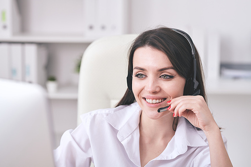 Young cheerful female customer support representative with headset looking at computer screen while consulting clients online