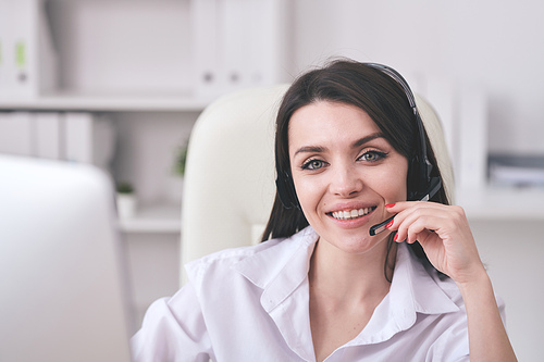 Portrait of smiling friendly helpdesk operator adjusting microphone headset while communicating with customer