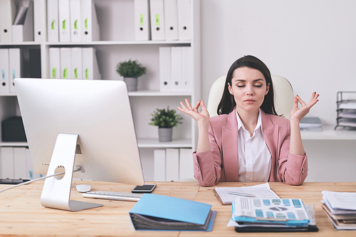 Young brunette businesswoman with closed eyes touching fingers while finding zen at workplace