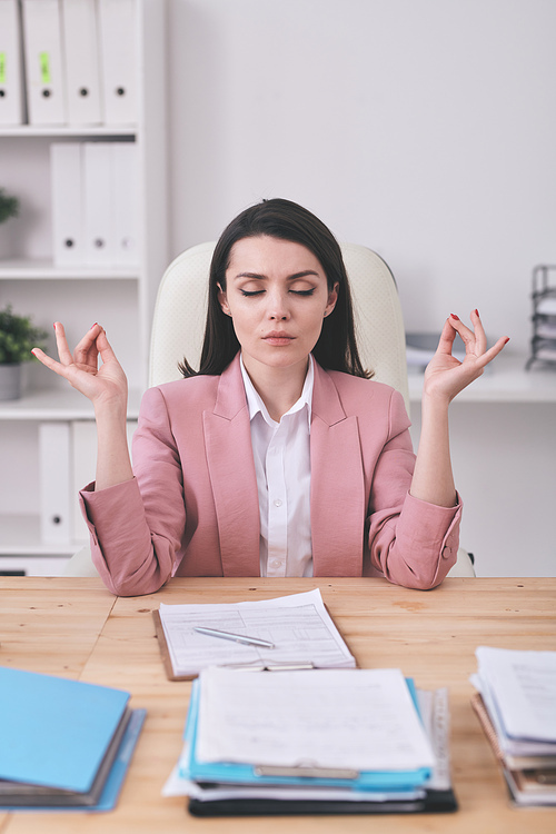 Young serious female office manager practicing yoga by desk while having break after work with financial documents or analyzing data