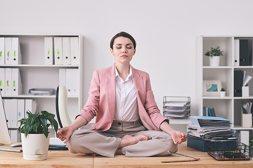 Young spiritual woman in pink jacket sitting in lotus position on table and meditating in office