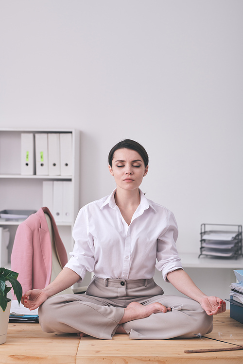calm young businesswoman with closed eyes sitting in zen- position and meditating in office