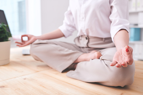 Close-up of unrecognizable businesswoman sitting in lotus position and holding hands in mudra while practicing relaxing meditation at workplace