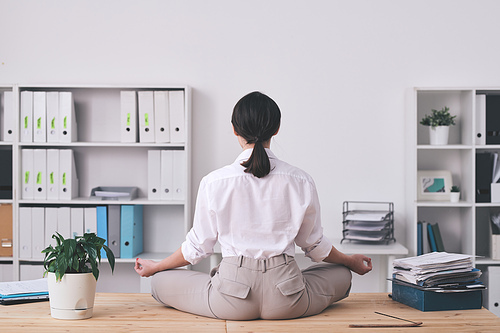 Rear view of brunette office woman sitting in lotus position on table and meditating in silence