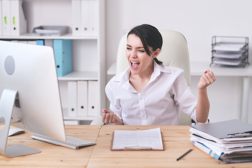 Jolly excited young female manager in white blouse looking at computer monitor and making yes gesture while winning tender
