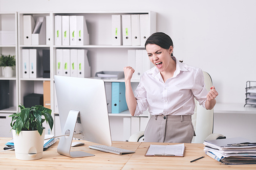 Young joyful businesswoman or female broker expressing triumph while standing by desk in front of computer monitor in office