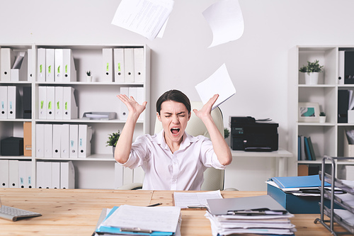 Frustrated young female manager annoyed with paperwork throwing papers and screaming with closed eyes in office