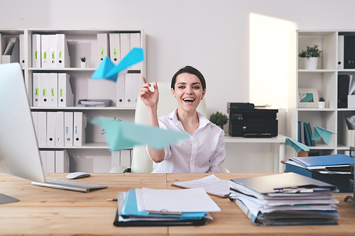 Cheerful excited young businesswoman sitting at desk full of documents and throwing paper plane in office