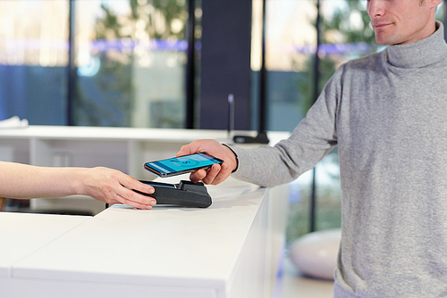 Young man in grey pullover holding smartphone with online banking page on screen during contactless payment process in shop or leisure center