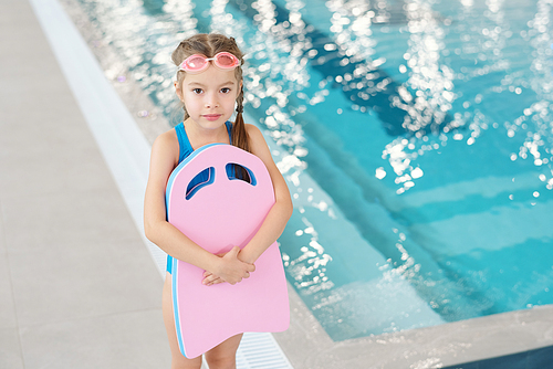 Adorable little girl in swimwear and goggles holding swimming board while standing in front of camera against poolside in sports center