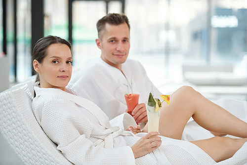 Young couple in white bathrobes relaxing in modern spa center and having tropical cocktails while sitting on deckchairs in front of camera