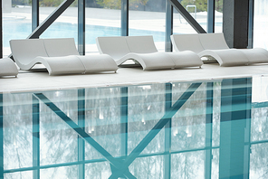 Row of white plastic deckchairs standing along large windows and swimming pool with transparent water inside modern spa center