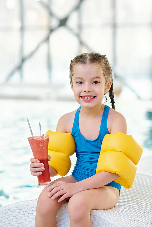 Cheerful little girl in swimwear and safety sleeves holding glass of juice or cocktail while sitting on deckchair in front of camera against poolside