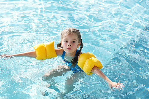 Blond little girl in blue swimwear and yellow safety sleeves on arms keeping balance in water of swimming pool and looking at you