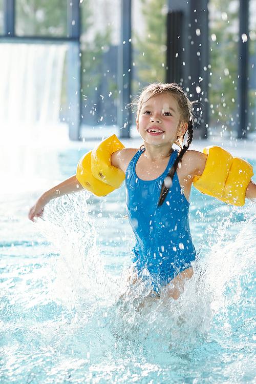 Laughing child in blue swimwear and yellow inflatable safety sleeves splashing water and having fun in swimming pool at leisure center