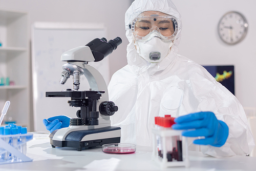 Young woman in coveralls, gloves, mask, and protective eyewear sitting in lab while working with samples of blood by microscope