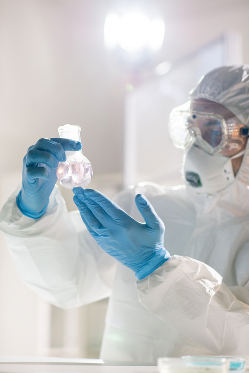 Laboratory worker or chemist in protective gloves, mask, eyeglasses and coveralls studying features of new liquid substance in tube