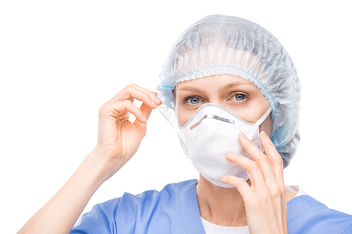 Portrait of blue-eyed woman in surgical cap putting respiratory mask on face against white background