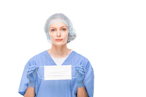Pretty young nurse or surgeon in blue uniform, gloves and headcloth holding protective mask in front of herself while standing in isolation
