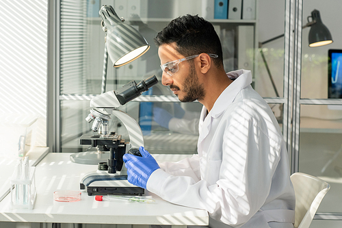 Side view of young male laboratory worker in gloves, eyeglasses and whitecoat studying sample of lab-grown soy sprout in microscope