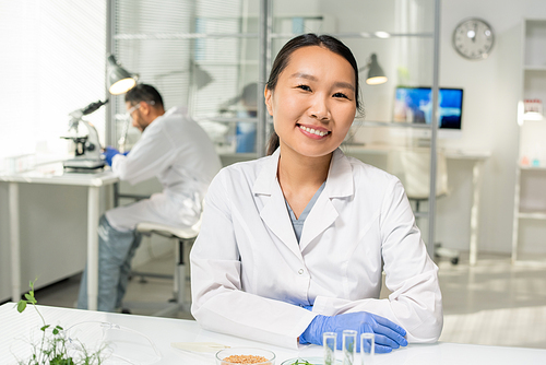 Young successful Asian female researcher in whitecoat and gloves looking at you with toothy smile while working with lab-grown sprouts